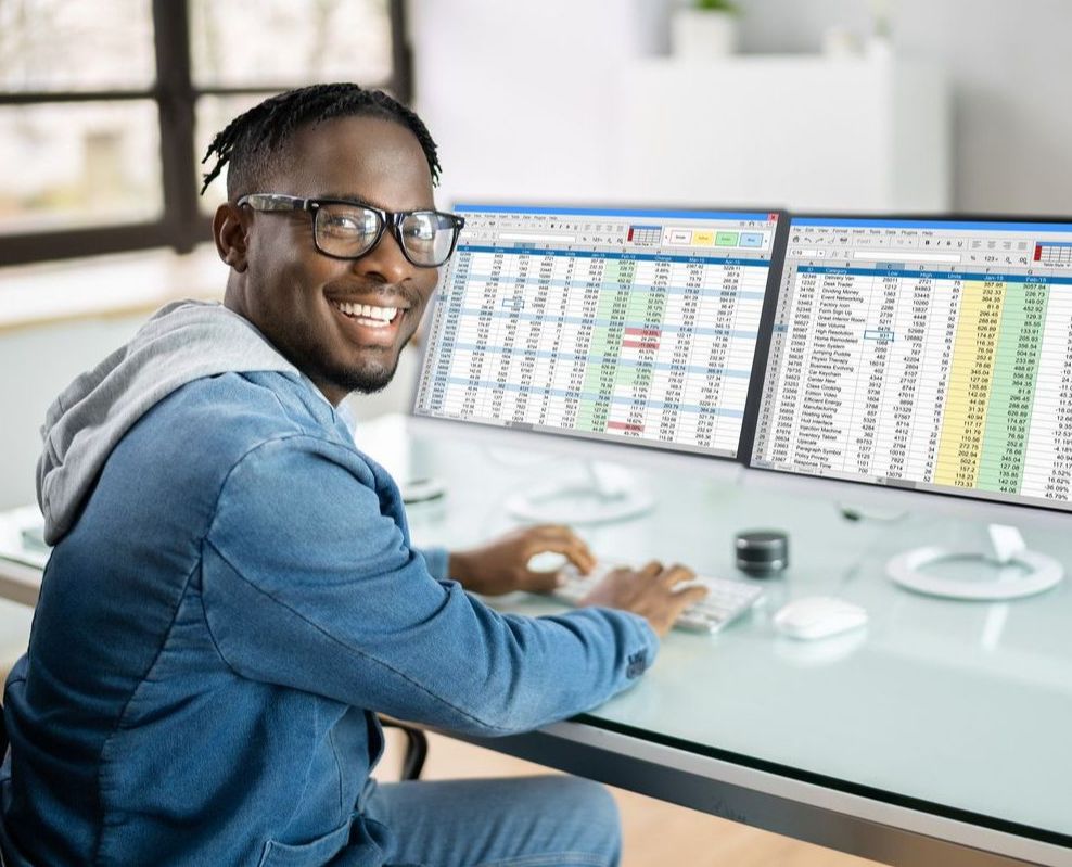 a man smiles while working at a desk with two computer monitors